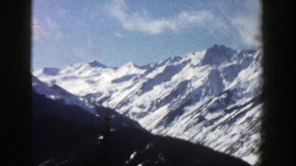 1961 Aspen, Colorado 8mm frame with jagged snowy peaks rising behind a dark lower ridge beneath scattered clouds.