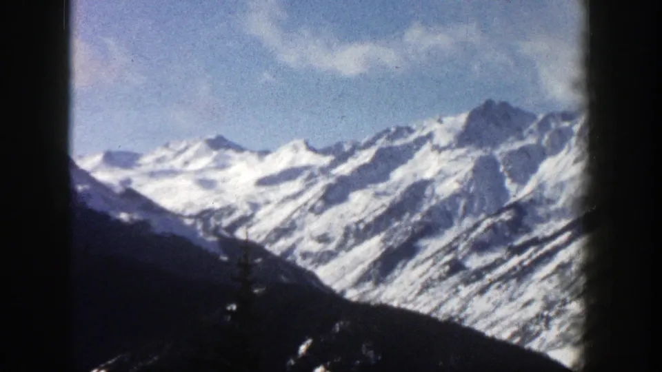 1961 Aspen, Colorado home-movie frame showing distant snowy Rockies ridges beneath blue sky and clouds, bordered by dark 8mm frame edges.