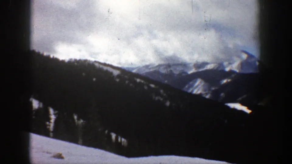 Vintage Aspen 8mm frame showing snow-streaked mountain ridges, dark wooded foreground, and a bright cloudy sky.