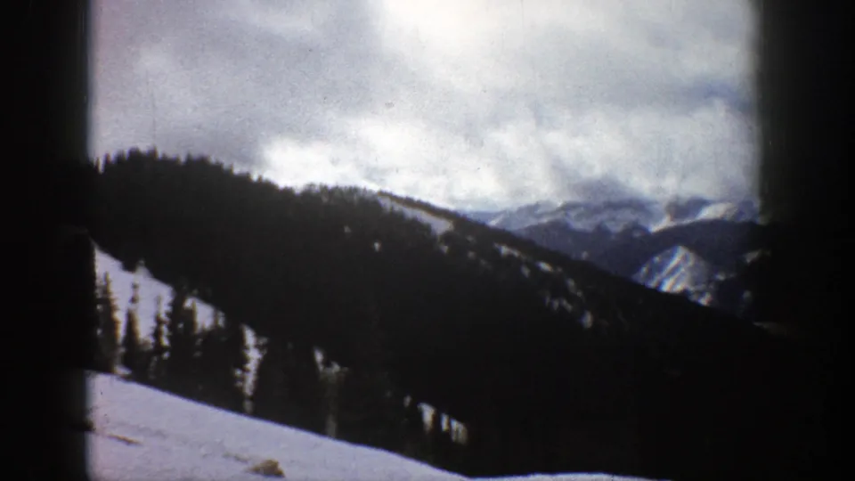 1961 Aspen, Colorado 8mm frame of a winter mountain vista with snowfields, evergreen-covered slopes, and soft distant peaks under clouds.