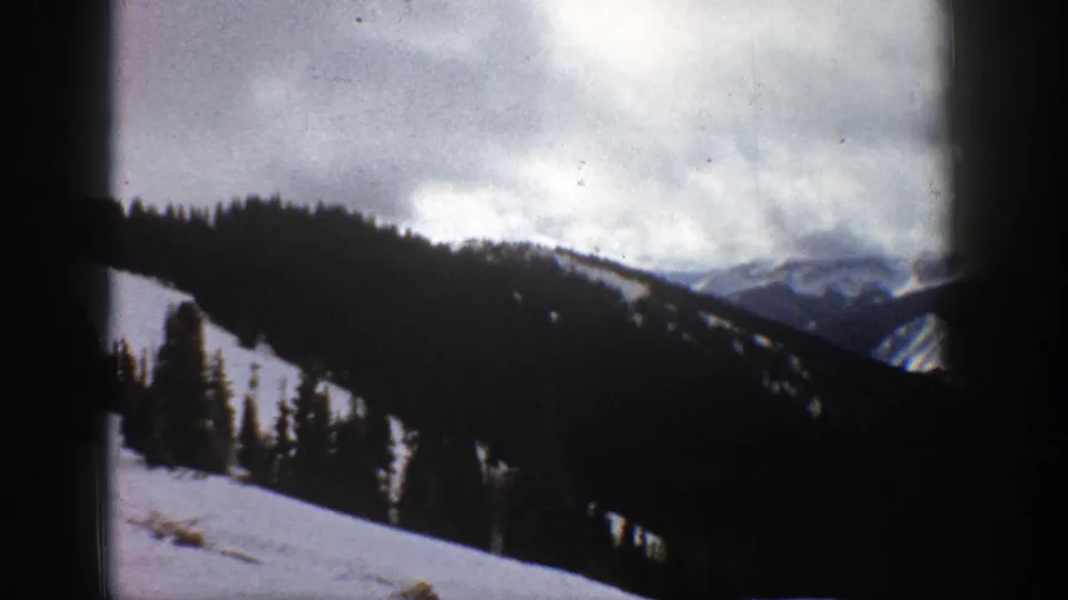1961 Aspen, Colorado 8mm frame with evergreen trees rising from a snowy slope and darker mountain ridges under a clouded sky.