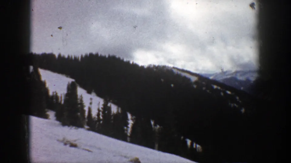 1961 Aspen, Colorado 8mm frame showing a snowy alpine hillside, scattered evergreen trees, distant ridges, and overcast mountain sky.