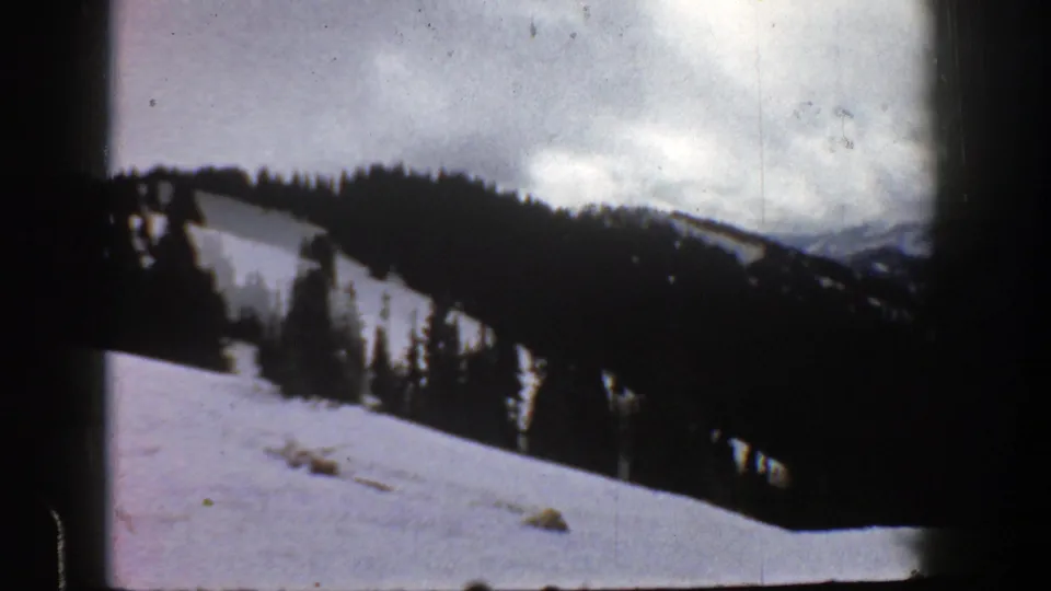 1961 Aspen 8mm frame showing a snow-covered mountain slope, evergreen tree line, distant ridges, and cloudy winter sky.