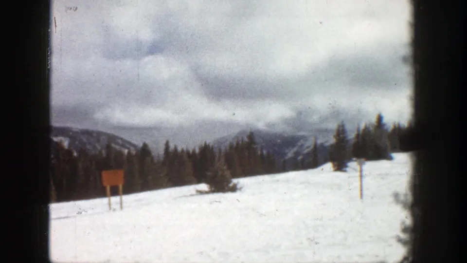 1961 Aspen 8mm frame with a wooden signpost standing in snow below evergreens and hazy mountain ridges.
