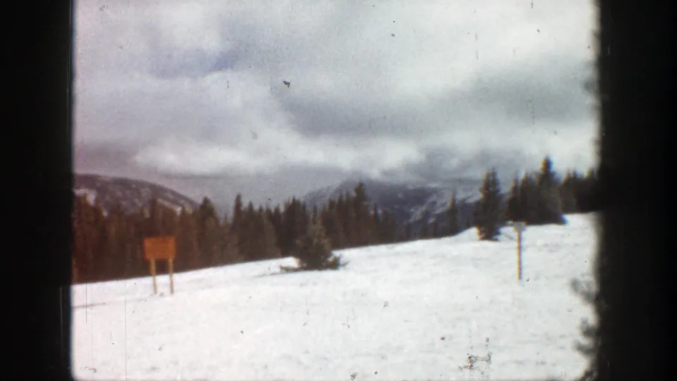 1961 Aspen 8mm frame showing a snow-covered clearing, unreadable wooden sign, scattered evergreens, and distant winter ridges.