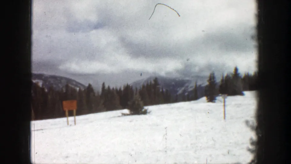 1961 Aspen 8mm frame of a snowy hillside with a wooden signpost, evergreen trees, distant mountains, and visible film-frame edges.