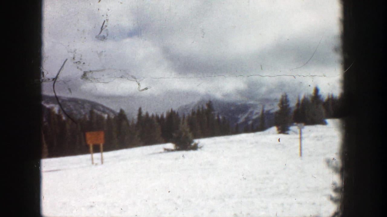 1961 Aspen home-movie frame showing a snowy slope, pine trees, distant mountains, and a small slope sign or marker.