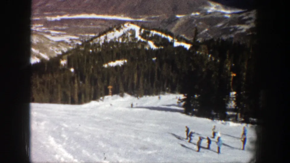 Vintage Aspen ski-slope frame showing small figures on snow with pine forest and rugged mountains behind them.