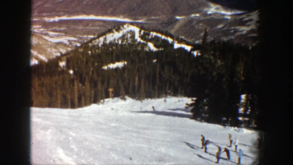 1961 Aspen home-movie still with tiny skiers on a bright slope, dark pines, and snow-streaked mountains.