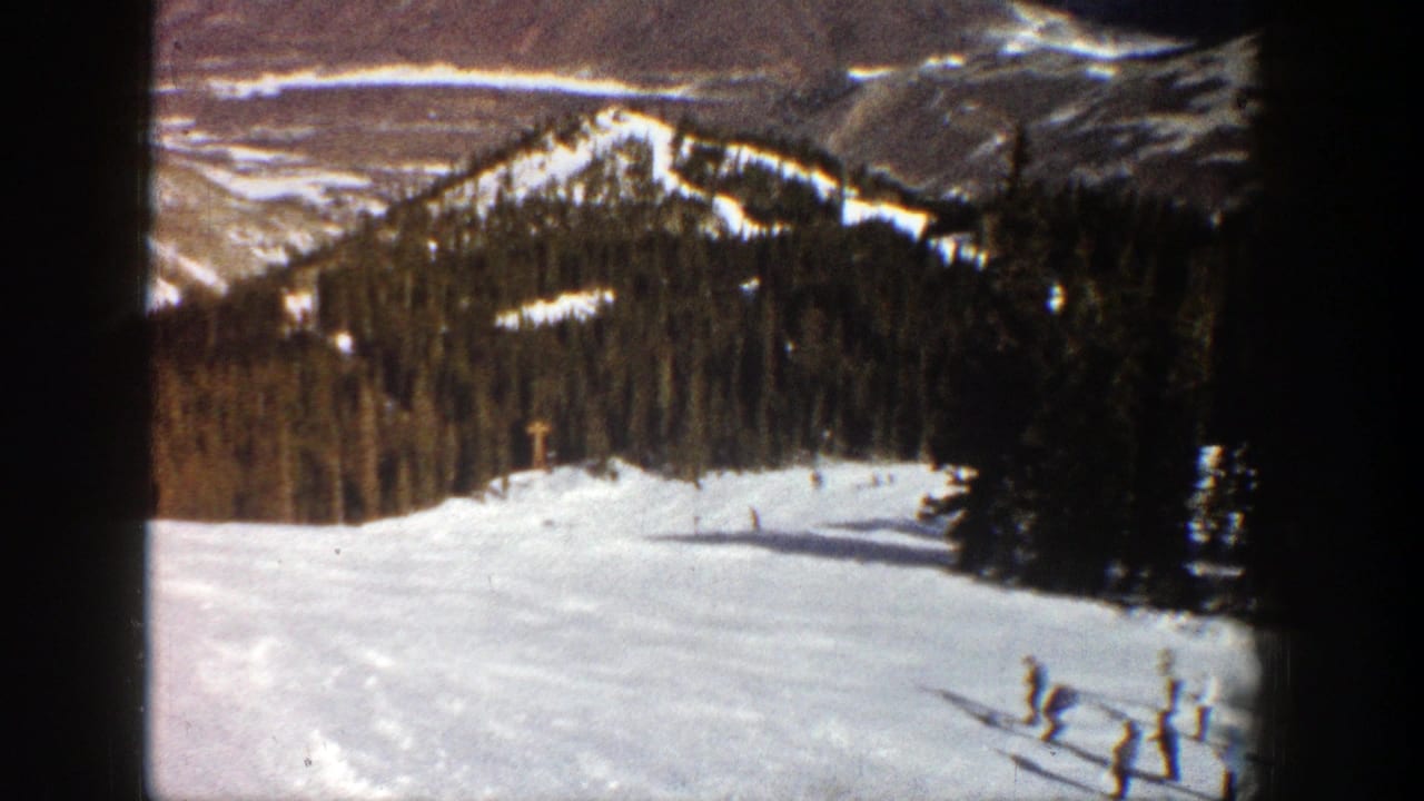 Skiers descend a snowy mountain run bordered by evergreen trees.