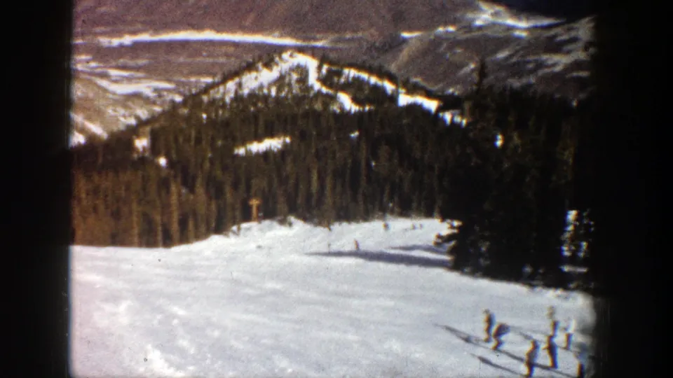 1961 Aspen home-movie frame showing skiers on a snow-covered slope with trees and mountain ridges beyond.