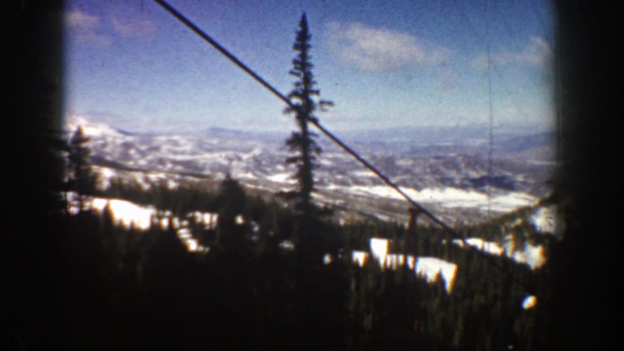 1961 8mm Aspen, Colorado view from a ski lift, with a cable cutting across blue sky above snowy ridges, evergreens, and a distant valley.
