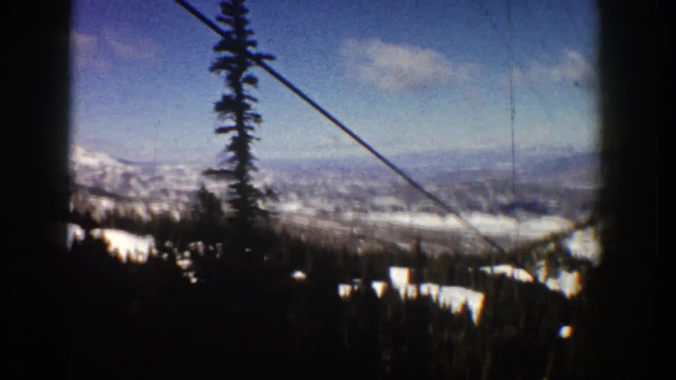 1961 8mm Aspen, Colorado frame looking past a tall evergreen and diagonal ski-lift cable toward snow patches, ridges, and a hazy valley.