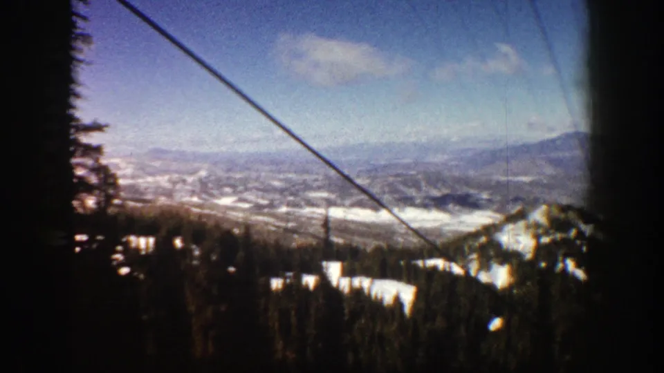 1961 8mm Aspen, Colorado ski-lift perspective across evergreens and snow-covered slopes toward a broad mountain valley under blue sky.