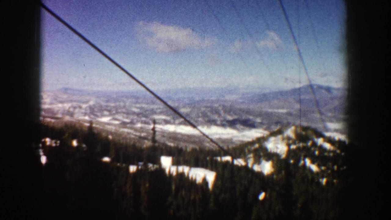 1961 Aspen, Colorado home-movie frame showing snowy mountain ridges, evergreen slopes, blue sky, and dark lift or gondola cables.