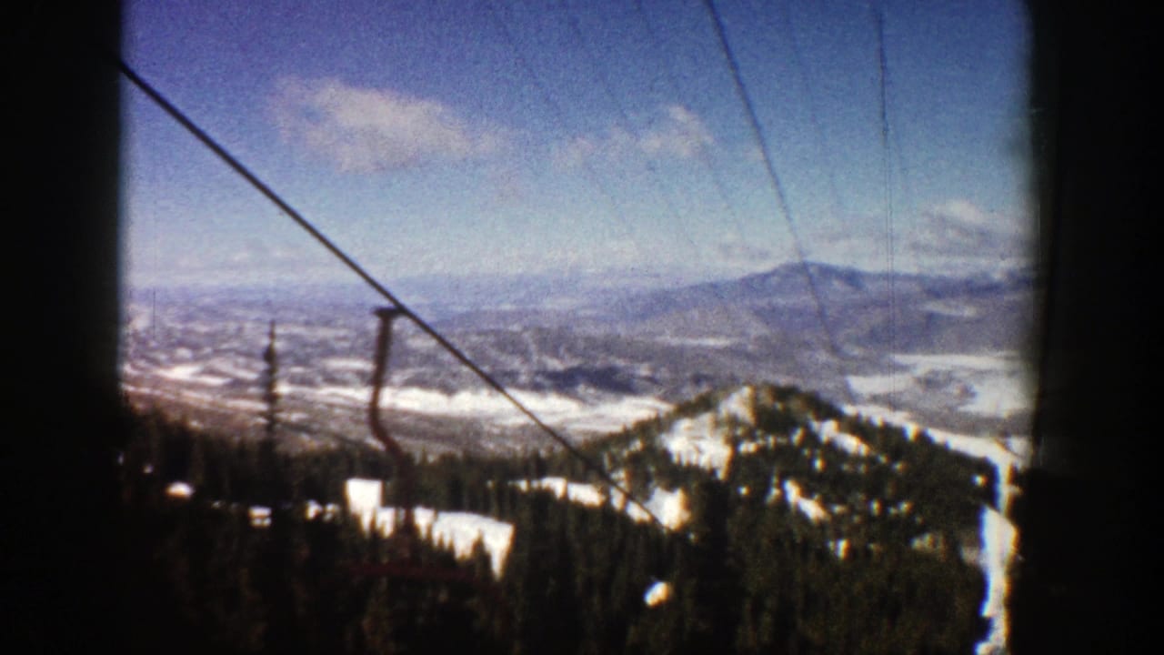 Mountain valley panorama with snow patches, evergreen slopes, and cables in the foreground.