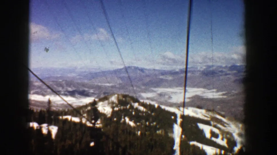 1961 Aspen home-movie frame with snowy ridge, distant valley, evergreen forest, blue sky, and lift or gondola cables in front.
