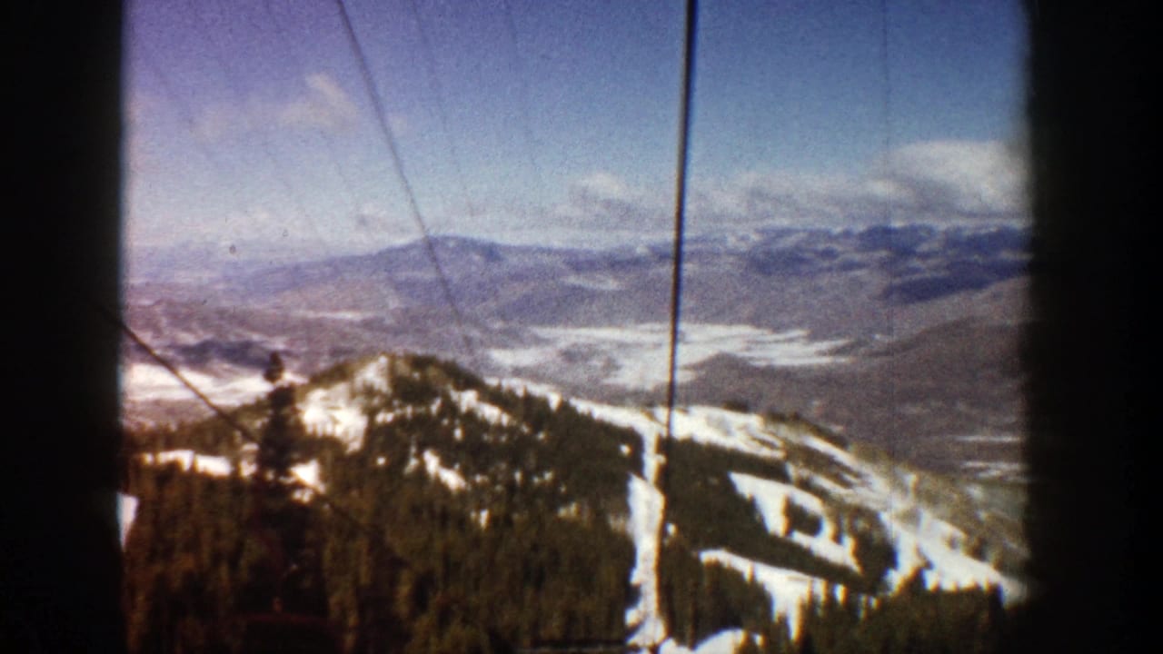Snowy mountain slope with evergreen trees, ski trails, distant valley, and lift cables in frame.