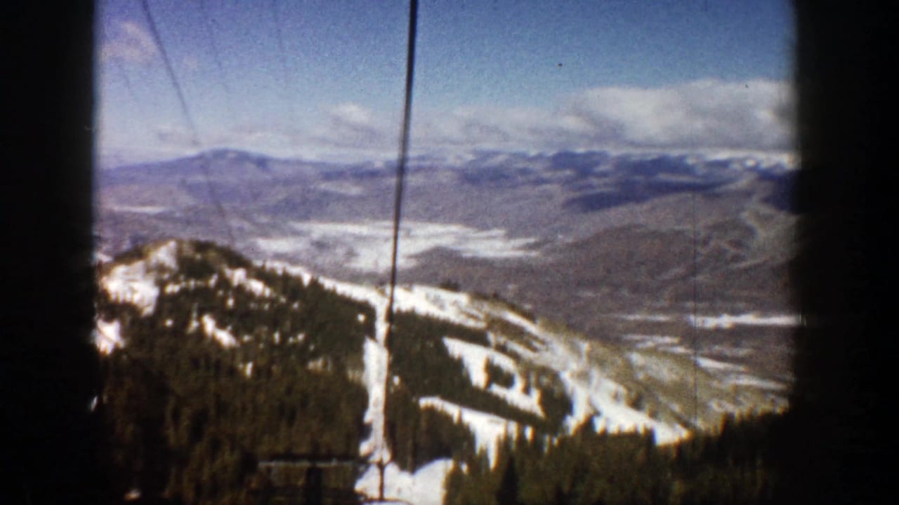 Snowy forested ridge with winding ski trails, distant mountains, clouds, and vertical lift lines.