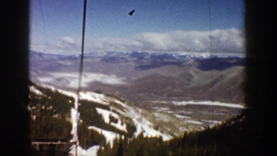 1961 8mm frame looking from an Aspen ski lift toward distant ridges and a valley, with snowy trails at the lower edge.