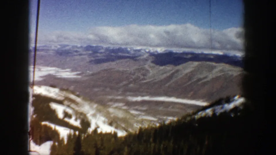 Aspen ski-lift home-movie frame showing a wide snowy mountain panorama, evergreen slope, and cloudy horizon.