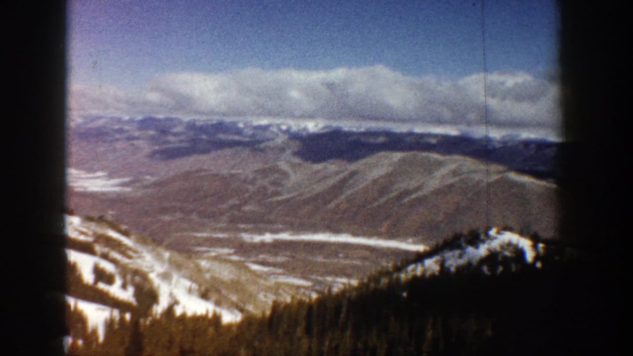 1961 Aspen home-movie frame showing a snow-dusted mountain valley, dark evergreen trees, and a cloudy blue sky.