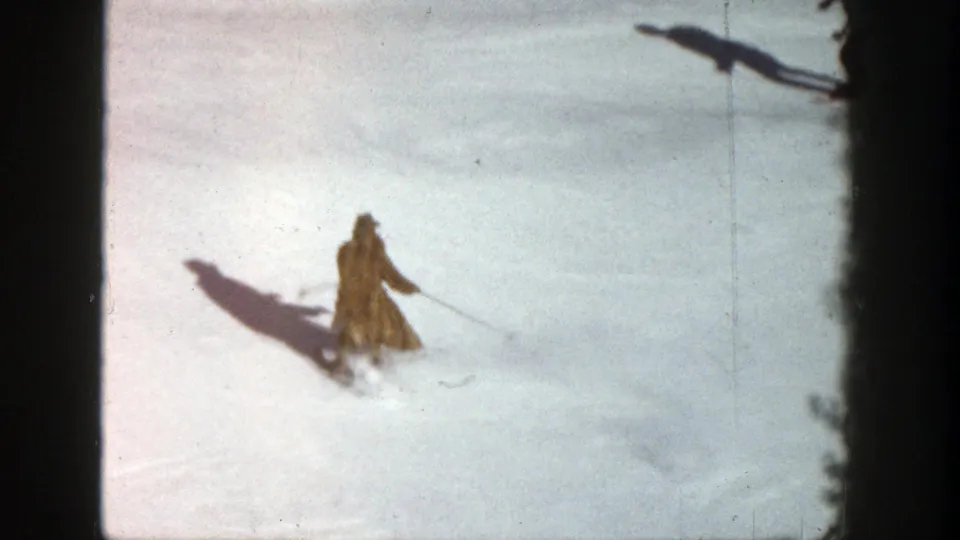 1961 Aspen frame of a brown-coated skier continuing downhill on sunlit snow near dark tree framing.
