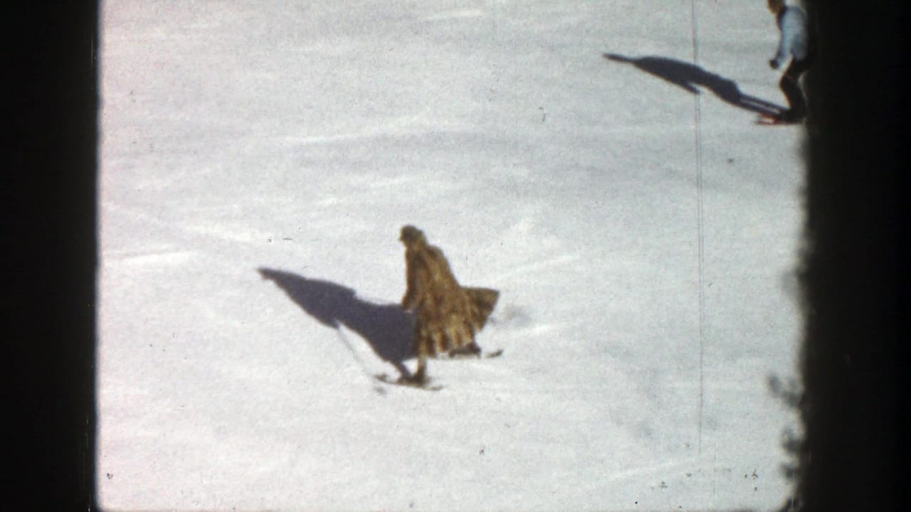 Skier in a long coat moving across bright snow with another skier partly visible above.