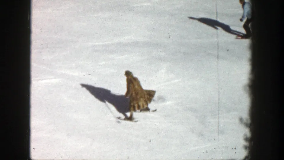 1961 Aspen home-movie frame showing a skier in a long patterned coat crossing a snowy slope with another skier partly visible near the top edge.