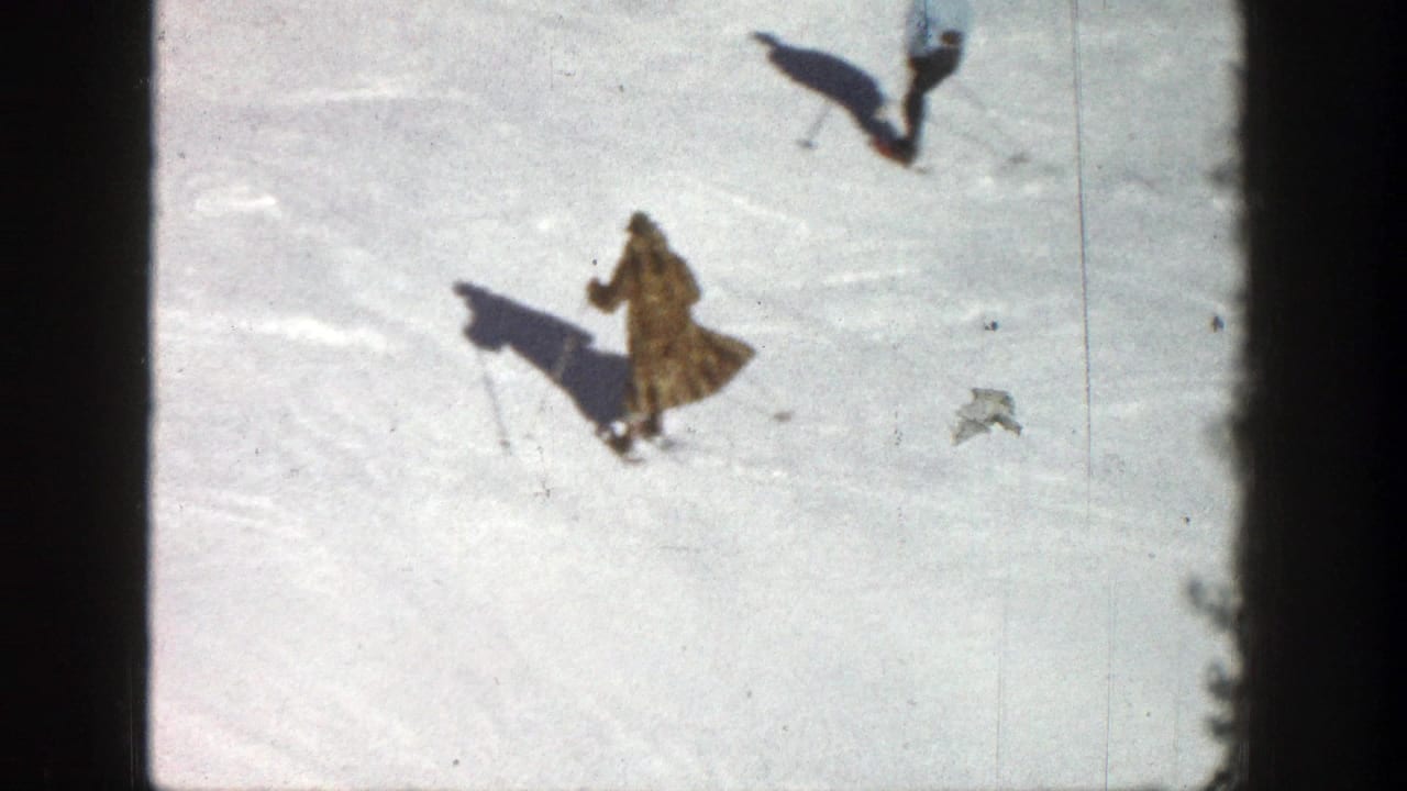 Two skiers cross a bright snowy slope, with one in a long coat below.
