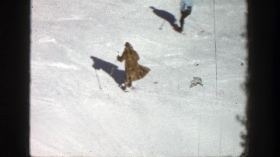 1961 Aspen skiing footage frame with two distant skiers on a sunlit snowfield, including a foreground skier in a long coat.
