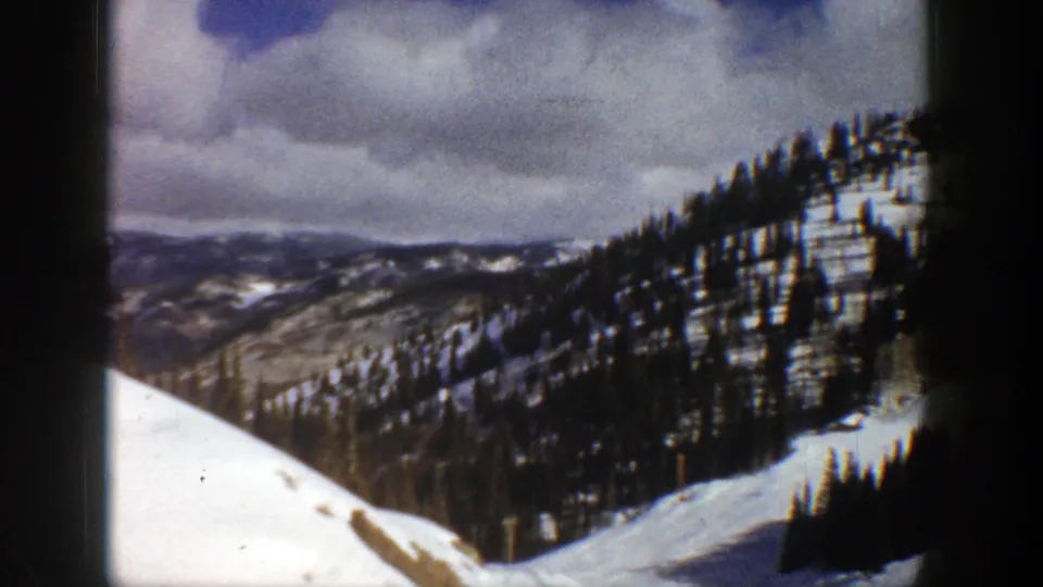 1961 Aspen, Colorado frame showing a snow-lined evergreen ridge with distant mountain terrain under overcast sky.