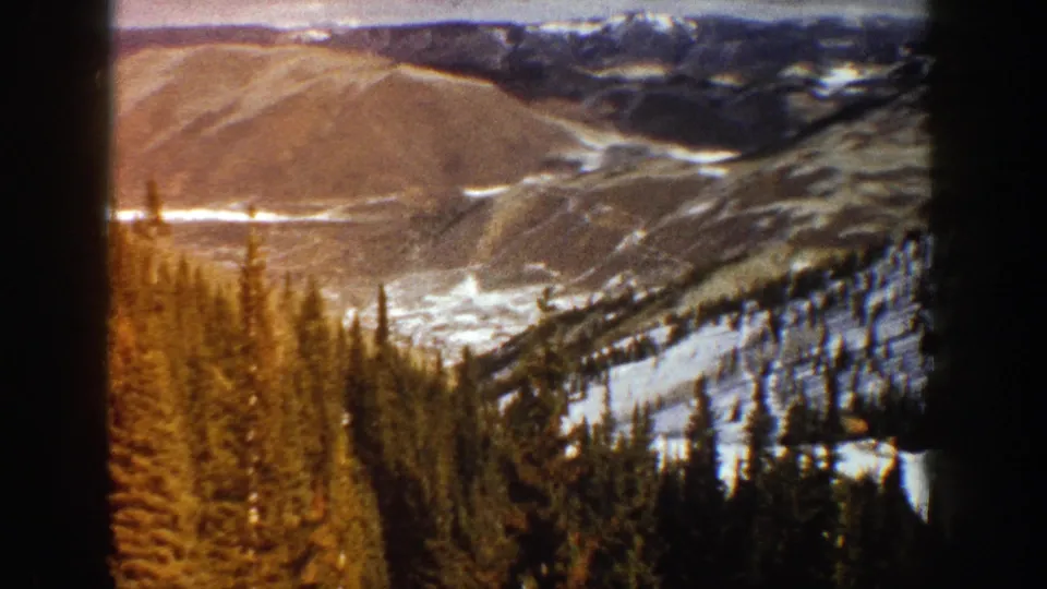 1961 Aspen-area home-movie frame showing dark evergreen forest below snow-dusted mountain slopes and distant ridgelines.