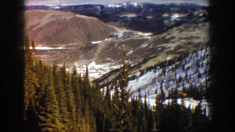 1961 8mm frame from Aspen showing a broad snowy valley, conifer forest, and muted mountain ridges under daylight.