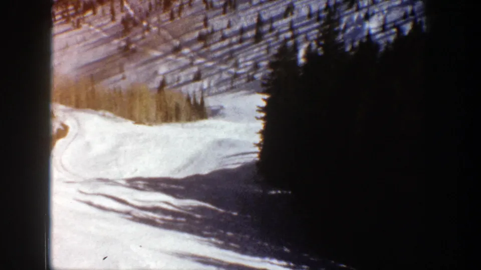 Home-movie view of an Aspen snow slope with strong evergreen shadows and warm-toned trees above the valley.