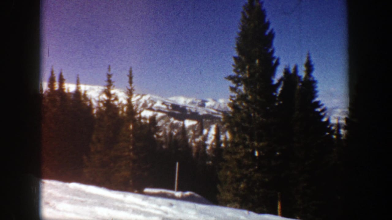 1961 Aspen-area winter view with tall evergreens, snow in the foreground, blue sky, and distant mountain ridges.