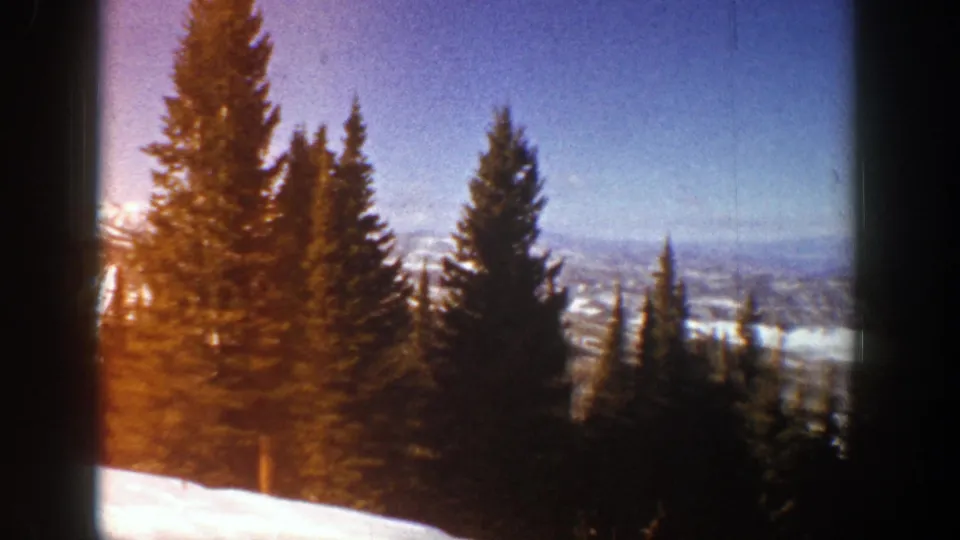 1961 Aspen, Colorado home-movie frame with evergreens silhouetted against snowy mountains and winter sky.