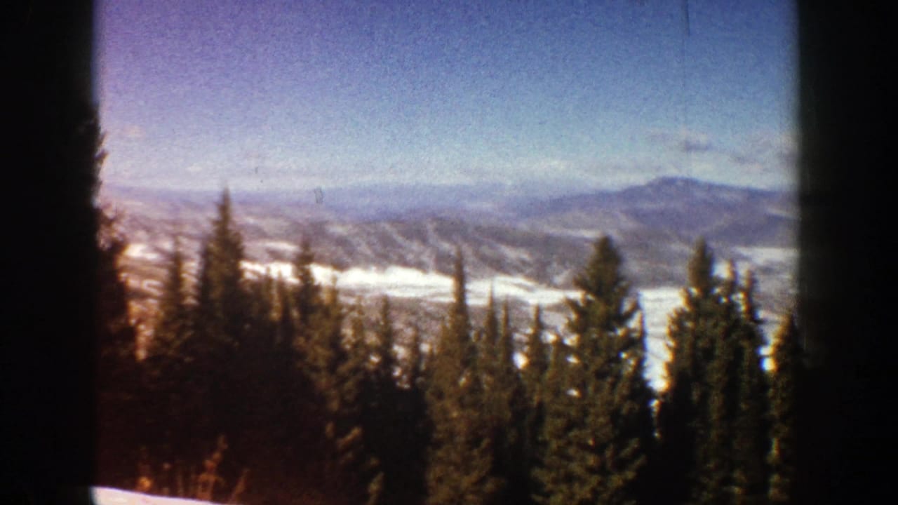 1961 Aspen, Colorado home-movie frame looking past evergreens toward a snowy mountain valley under pale blue sky.