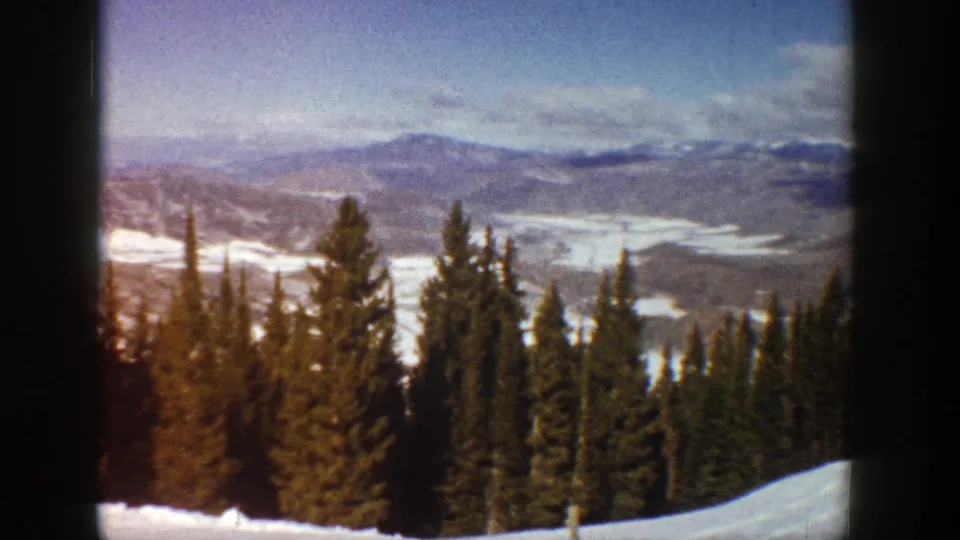 1961 Aspen, Colorado frame showing dark evergreens, a snowy foreground slope, and layered mountain ridges beyond.