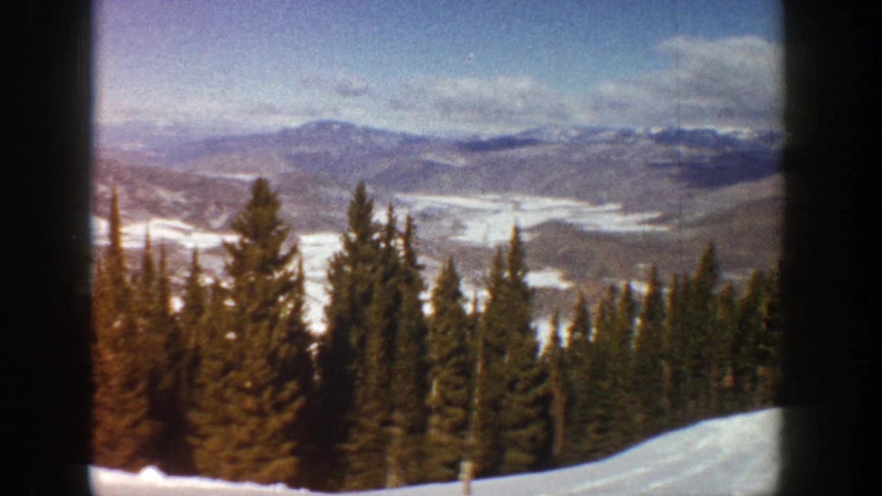 Snowy mountain overlook with evergreen trees and distant ridges under a cloudy sky.