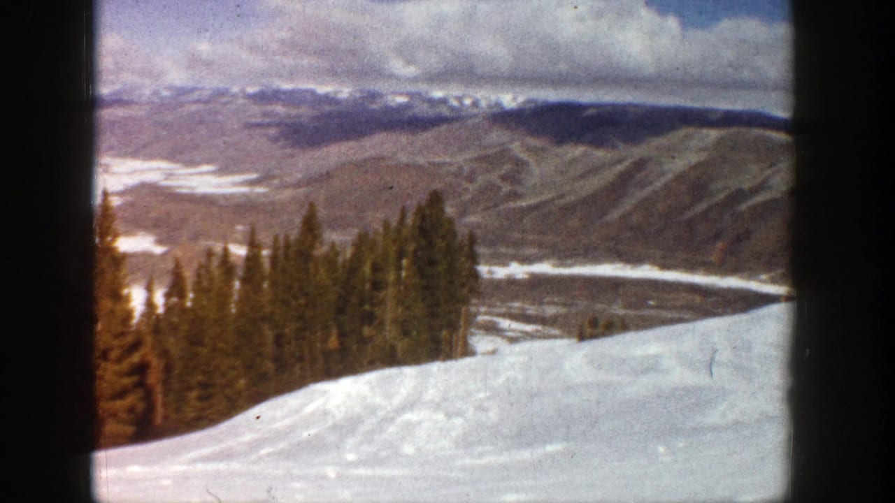 Snowy mountain slope with evergreen trees and distant ridgelines under clouds.