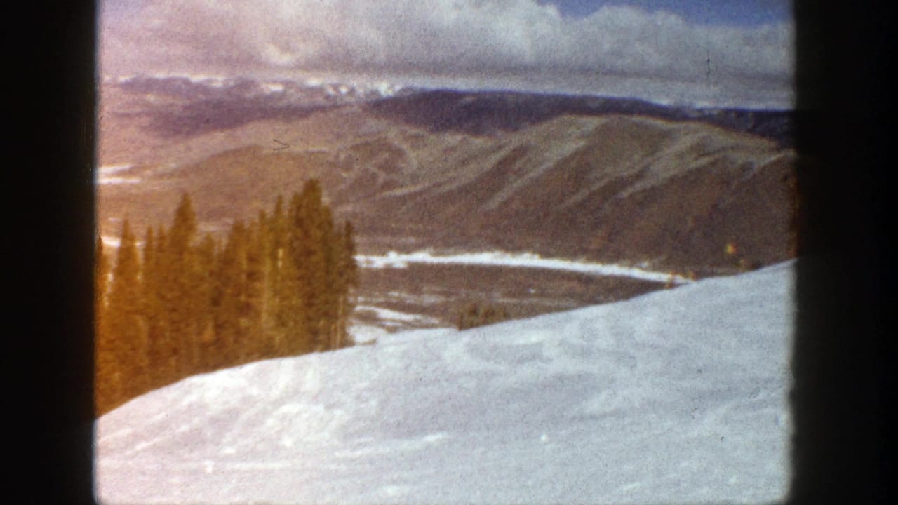 Snowfield and evergreen stand overlooking distant mountain ridges beneath clouds.