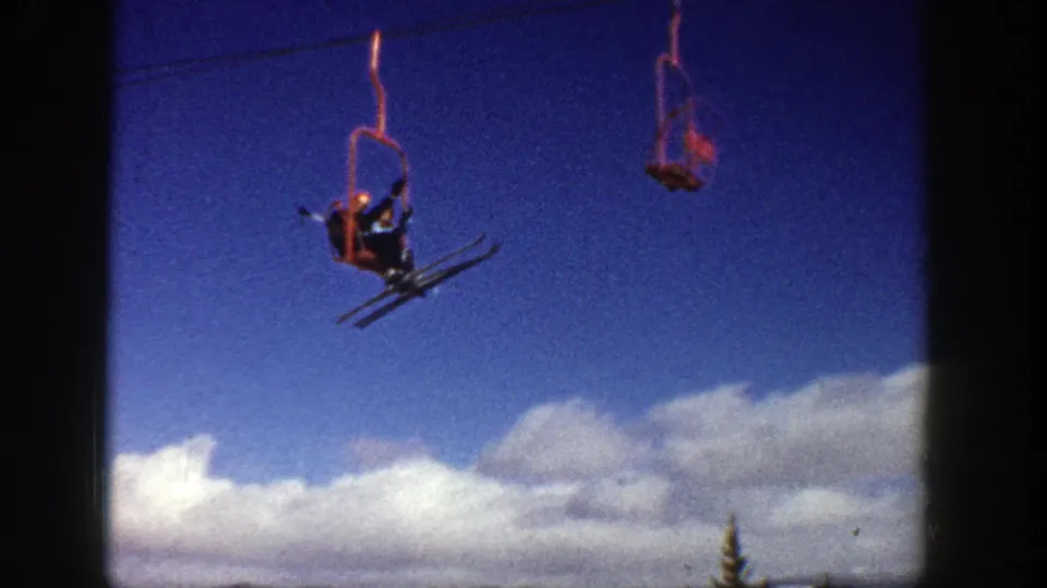 1961 Aspen 8mm frame showing one distant skier on a red chairlift, skis visible, suspended against blue sky with clouds and treetops below.