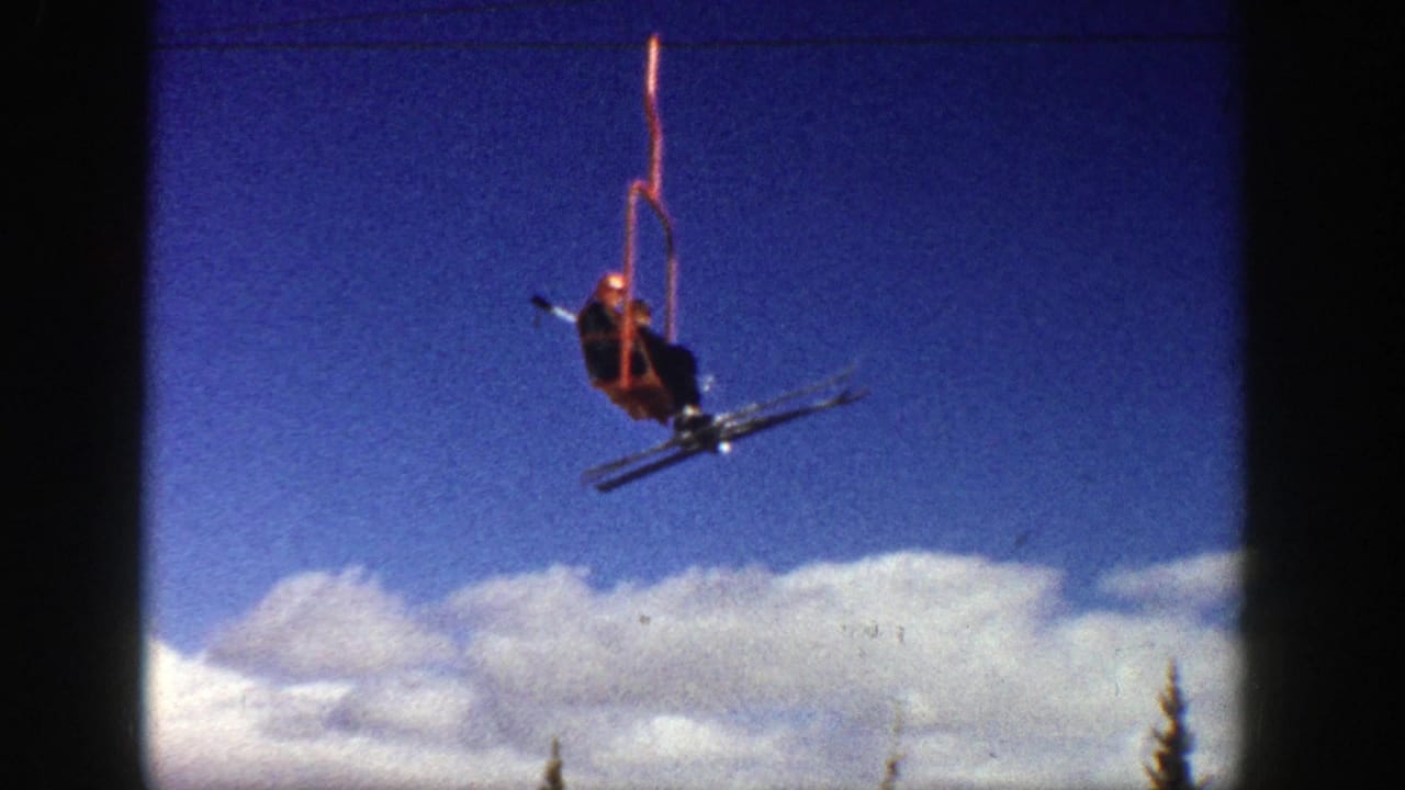 Skier on a red chairlift moves across the sky above clouds and snow.