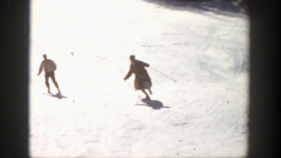 Two skiers move downhill across a highly sunlit snowfield, their figures softened by the 8mm home-movie exposure in the 1961 Aspen scene.