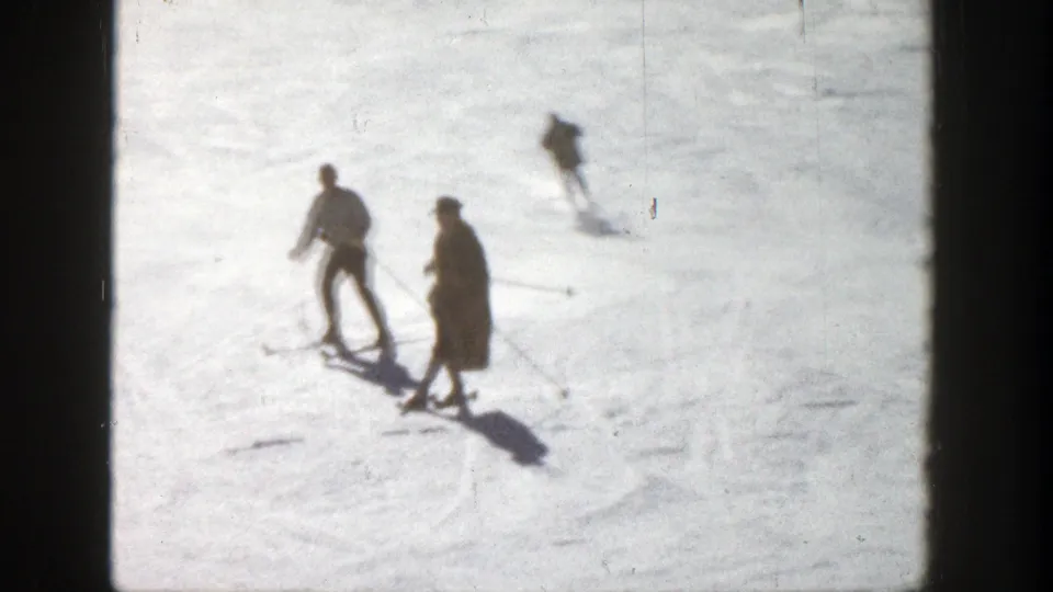 Multiple skiers travel across the snow, including two closer figures with poles and another skier farther uphill, in the supplied Aspen 1961 ski footage.