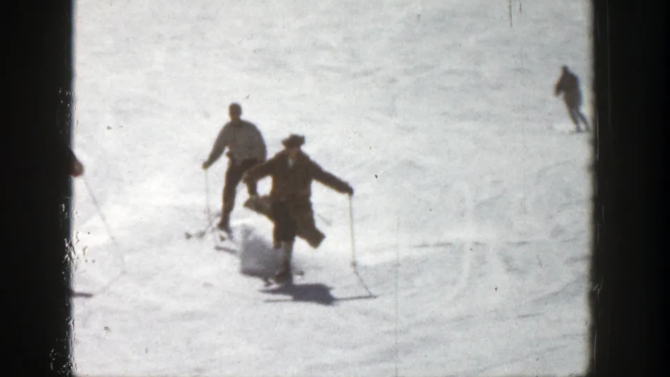 In 1961 Aspen home-movie footage, two skiers move downhill on bright snow as a distant figure crosses the slope behind them.