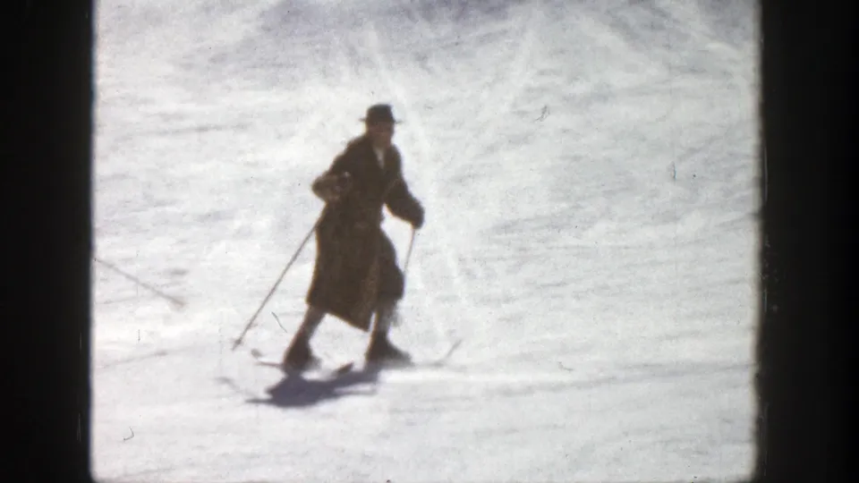 A single skier in a long coat and dark hat glides across the Aspen snow, captured with soft 8mm home-movie blur.