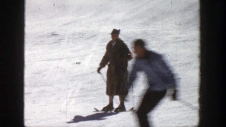 On the Aspen slope, one skier in a long coat pauses while another blurred skier passes close to the handheld camera.