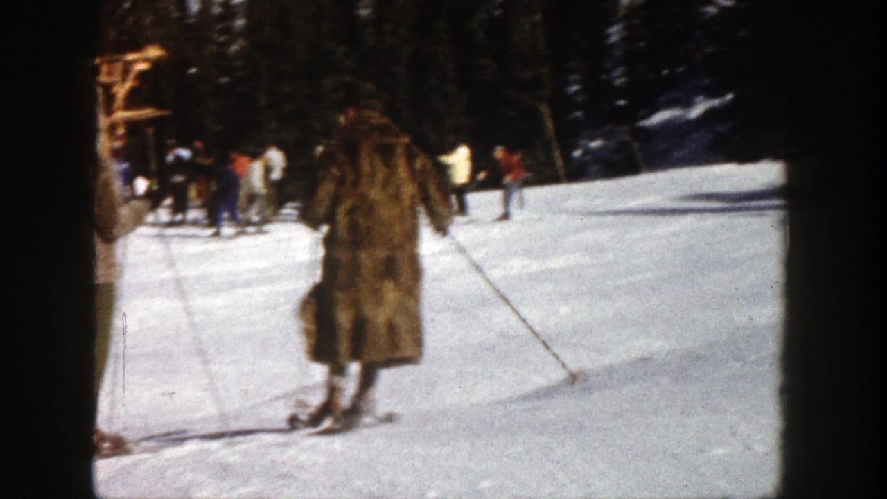 1961 Aspen home-movie frame showing a fur-coated skier on snow, with distant skiers, trees, and a slope-side structure behind.